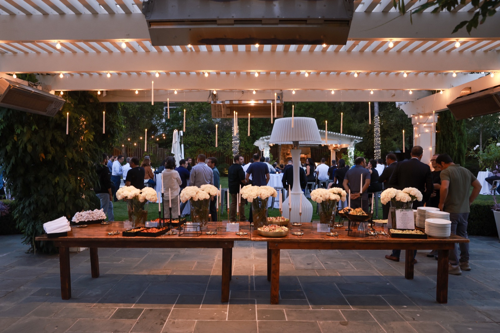 Outdoor evening cocktail event with string lights and white floral arrangements on wood tables