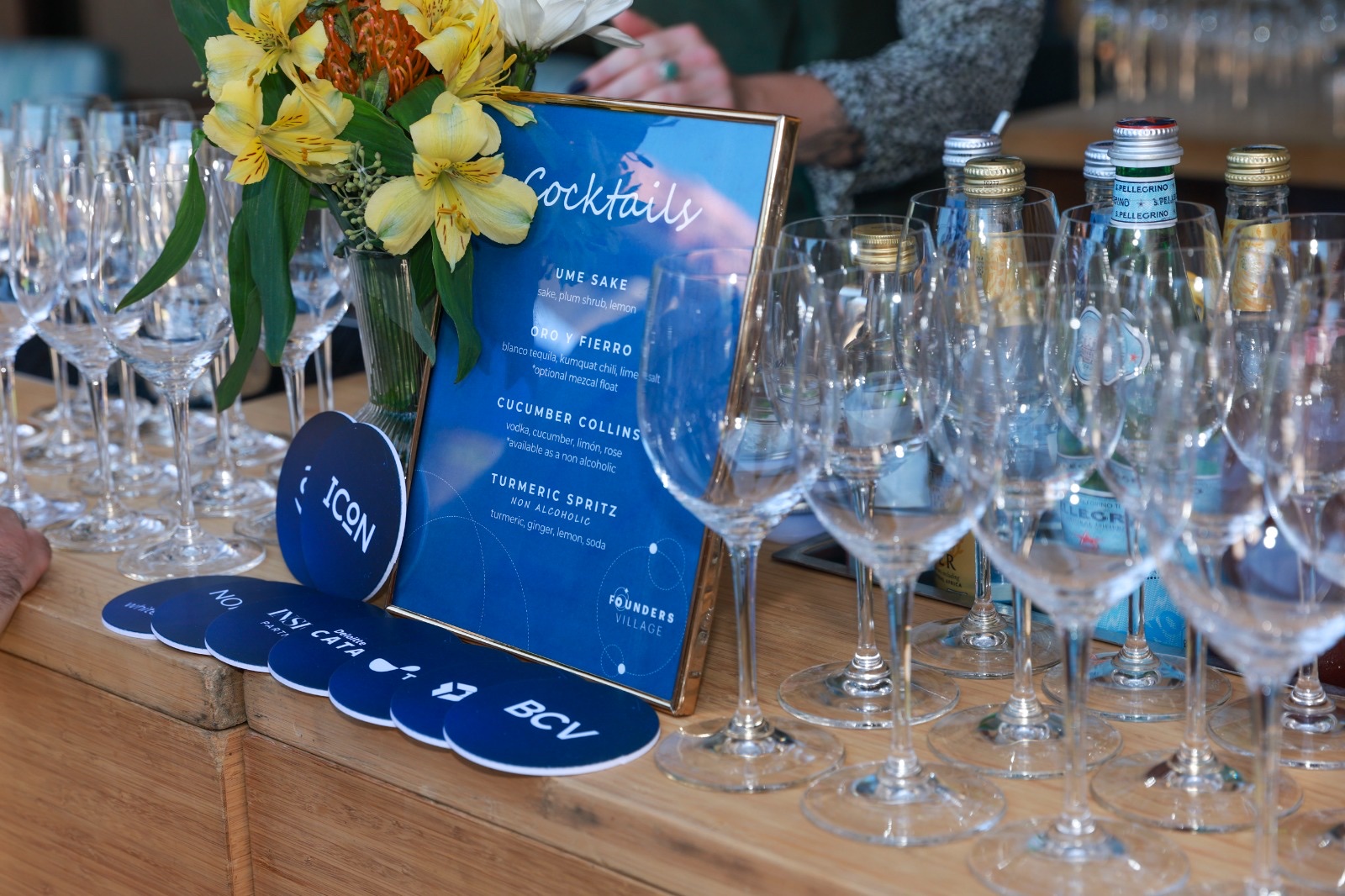 Cocktail menu sign reading 'Cocktails' in front of branded VC coasters and rows of wine glasses