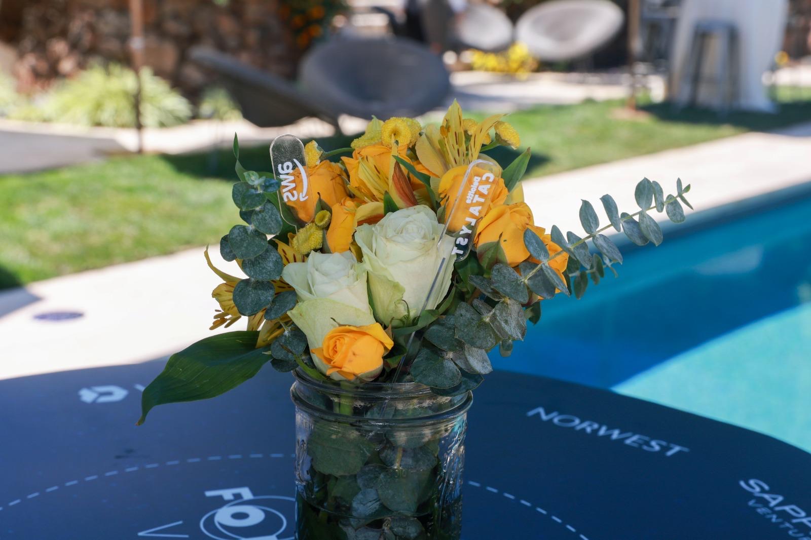 Yellow rose centerpiece in a mason jar with branded SMS and Catalyst stir-stick stakes by a pool