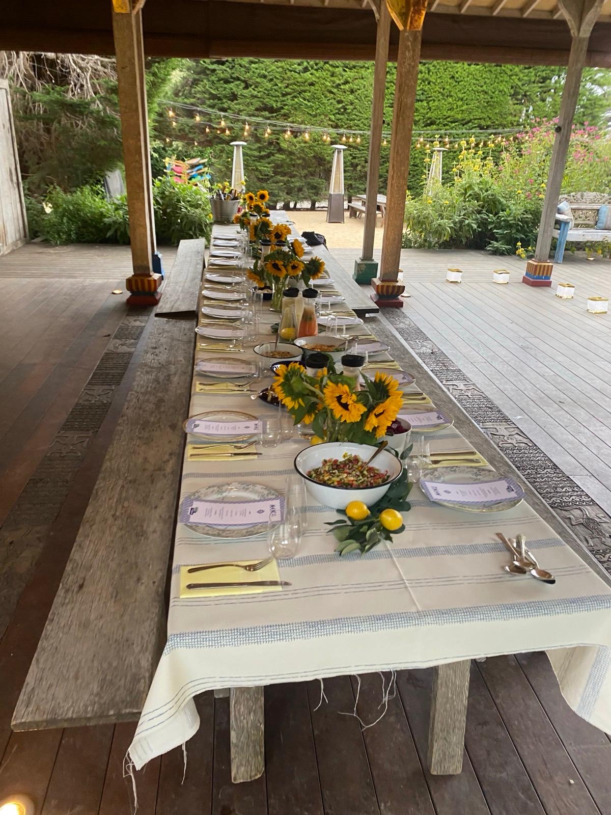 Long farm dinner table set with sunflowers, gold flatware, and string lights overhead